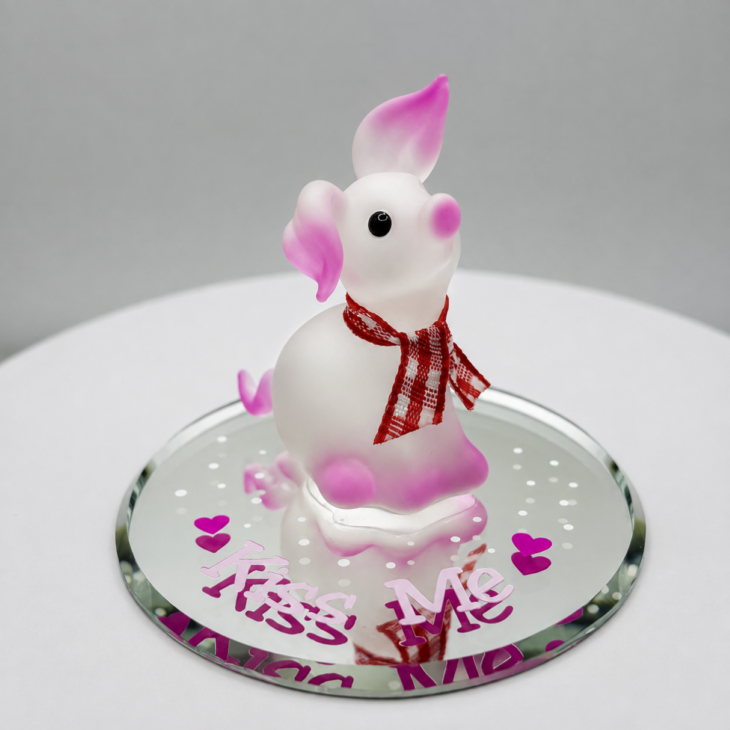 Handcrafted white and pink glass pig figurine with a red and white scarf on a beveled glass mirror engraved with "Kiss Me."