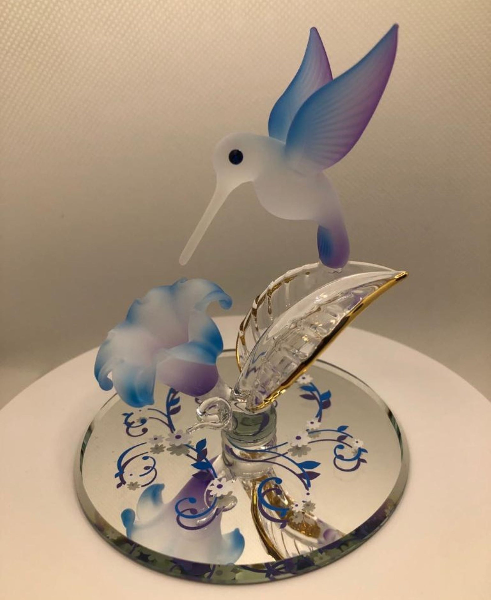 Handcrafted glass hummingbird poised over a blue flower with crystal and gold accents on a beveled mirror base