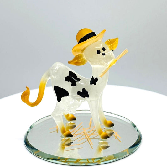 Handcrafted glass cow figurine with black spots and yellow hat on round glass display base